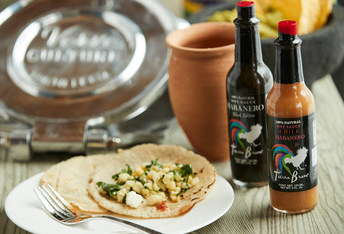 Two Tierra Brava habanero hot sauce bottles with red caps displayed next to a plated tostada and terracotta cup.