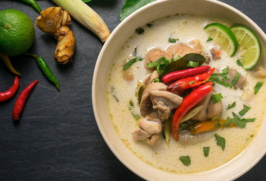 Creamy coconut soup with chicken, red chilies, lime slices, and herbs in a bowl with fresh ginger, lemongrass, and chilies ne