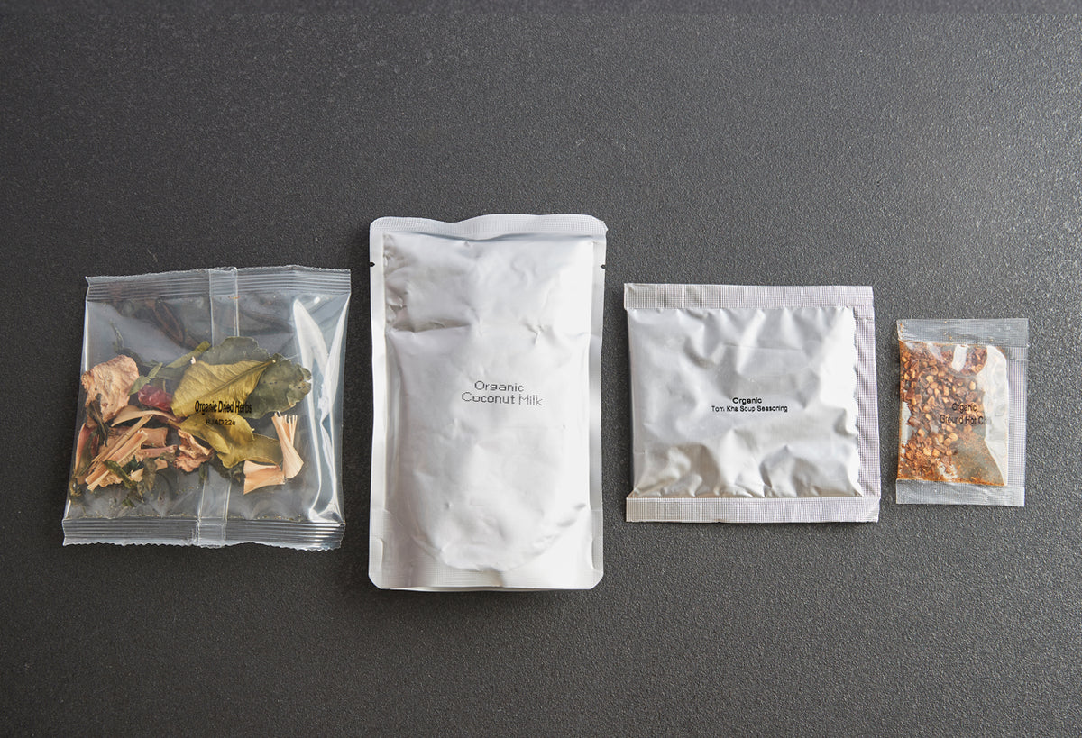 Four ingredient packets laid out on gray surface: dried herbs and spices in clear bag, organic coconut milk in white pouch, s