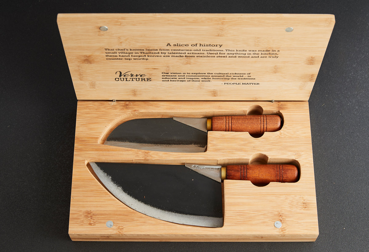 Two curved Thai chef knives with dark blades and wooden handles displayed in an open bamboo box with text.