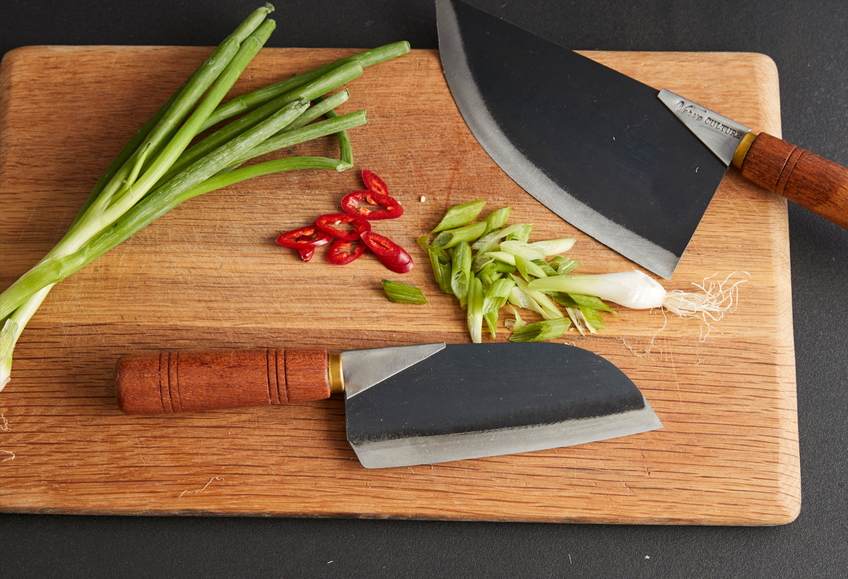 Two curved blade knives with wooden handles on cutting board with sliced green onions, red chilies, and green peppers.