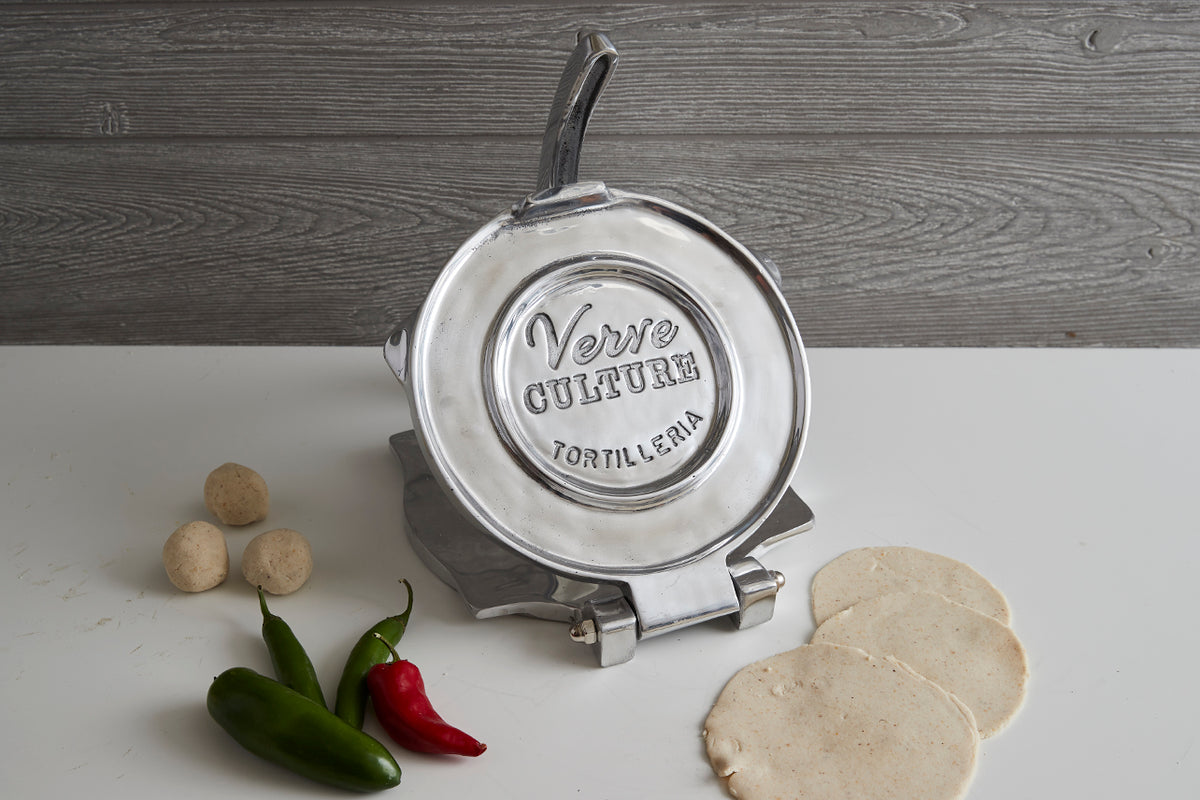 Polished aluminum tortilla press with black handle, displayed with two pressed tortillas, peppers, and dough balls on white s