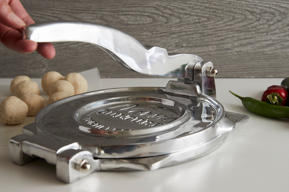 Polished aluminum tortilla press with handle being pressed down, potatoes and peppers nearby on white surface.