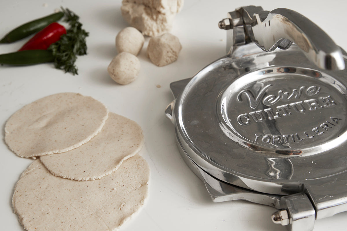 Polished aluminum tortilla press with handle next to three freshly pressed tortillas, dough balls, and fresh peppers.