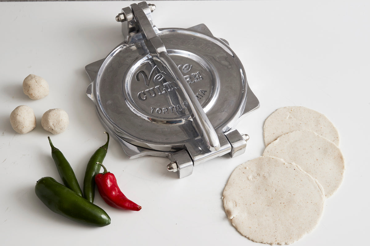 Polished aluminum tortilla press with lever handle, displayed with dough balls, pressed tortillas, and fresh chilies.