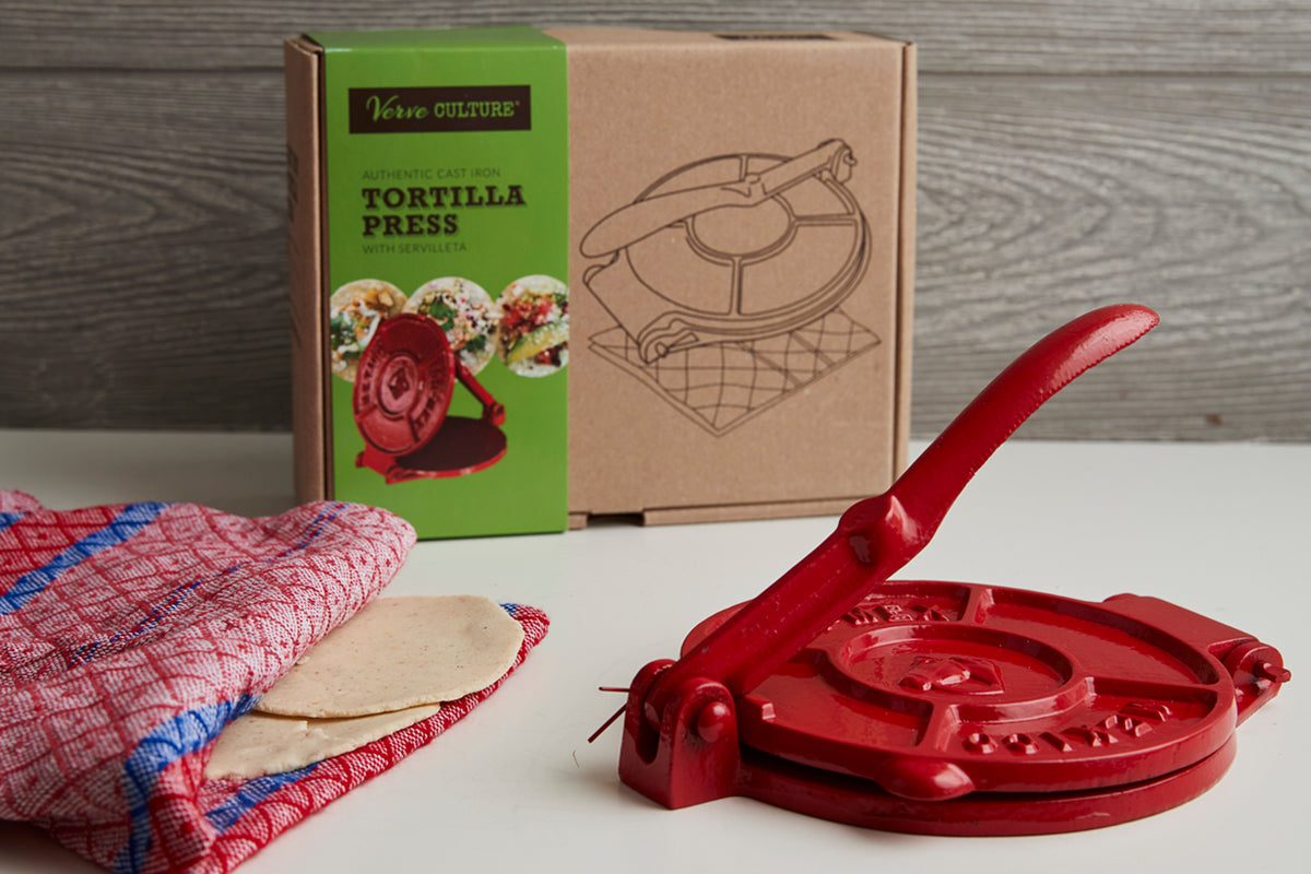 Red cast iron tortilla press with patterned cloth and boxed tortilla press packaging displayed on white surface.