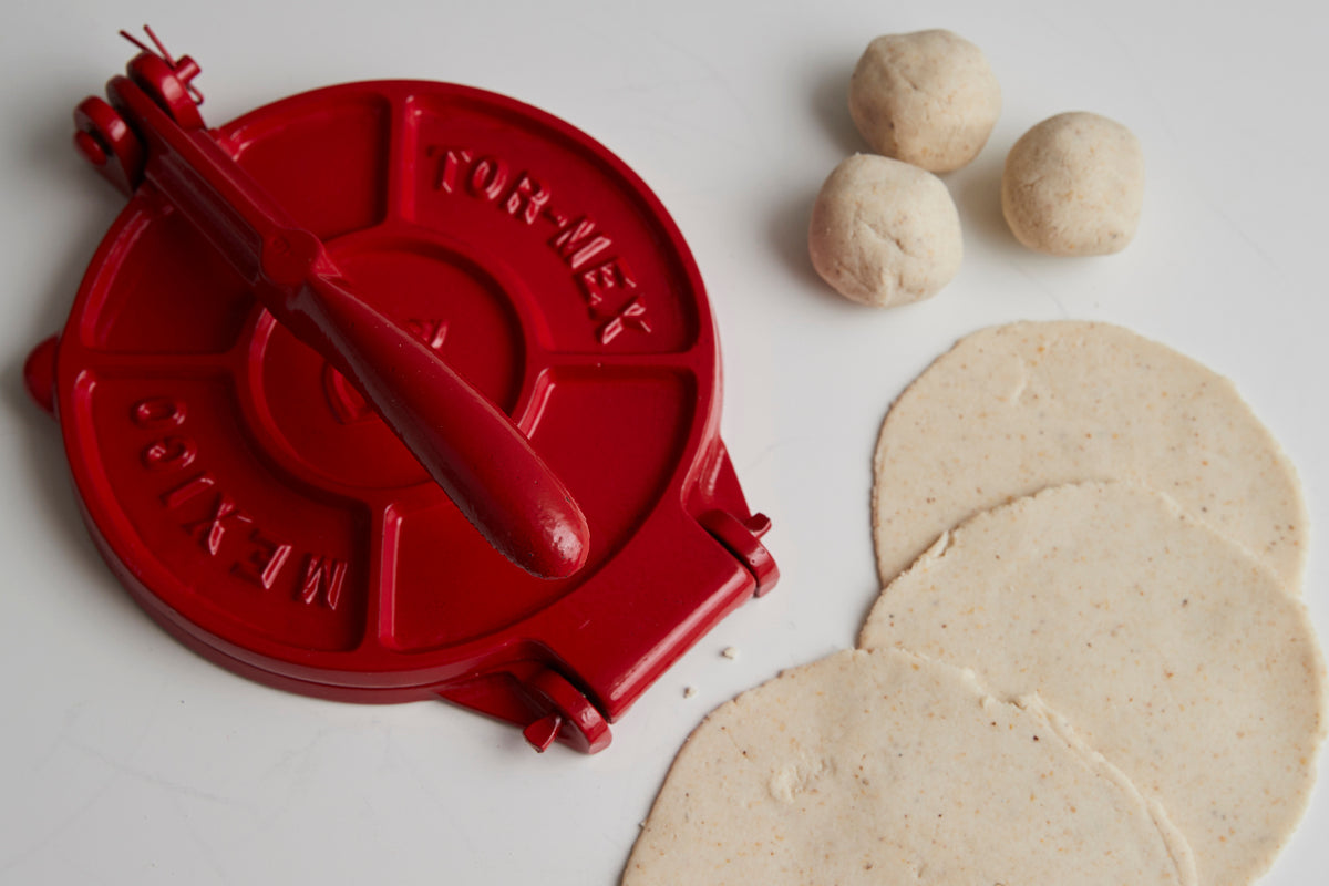 Red cast iron tortilla press with wooden handle, three dough balls, and two pressed tortillas.