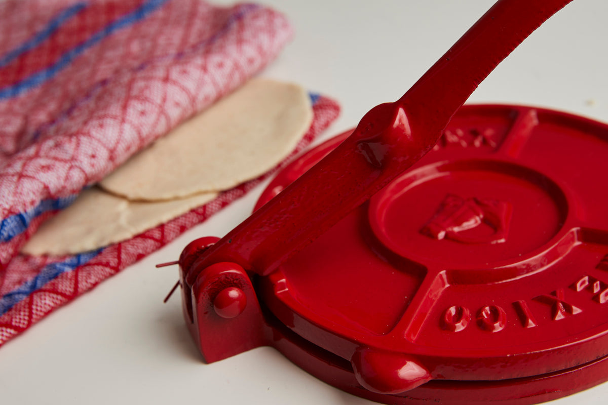 Red cast iron tortilla press with two pressed tortillas and red and blue striped cloth.