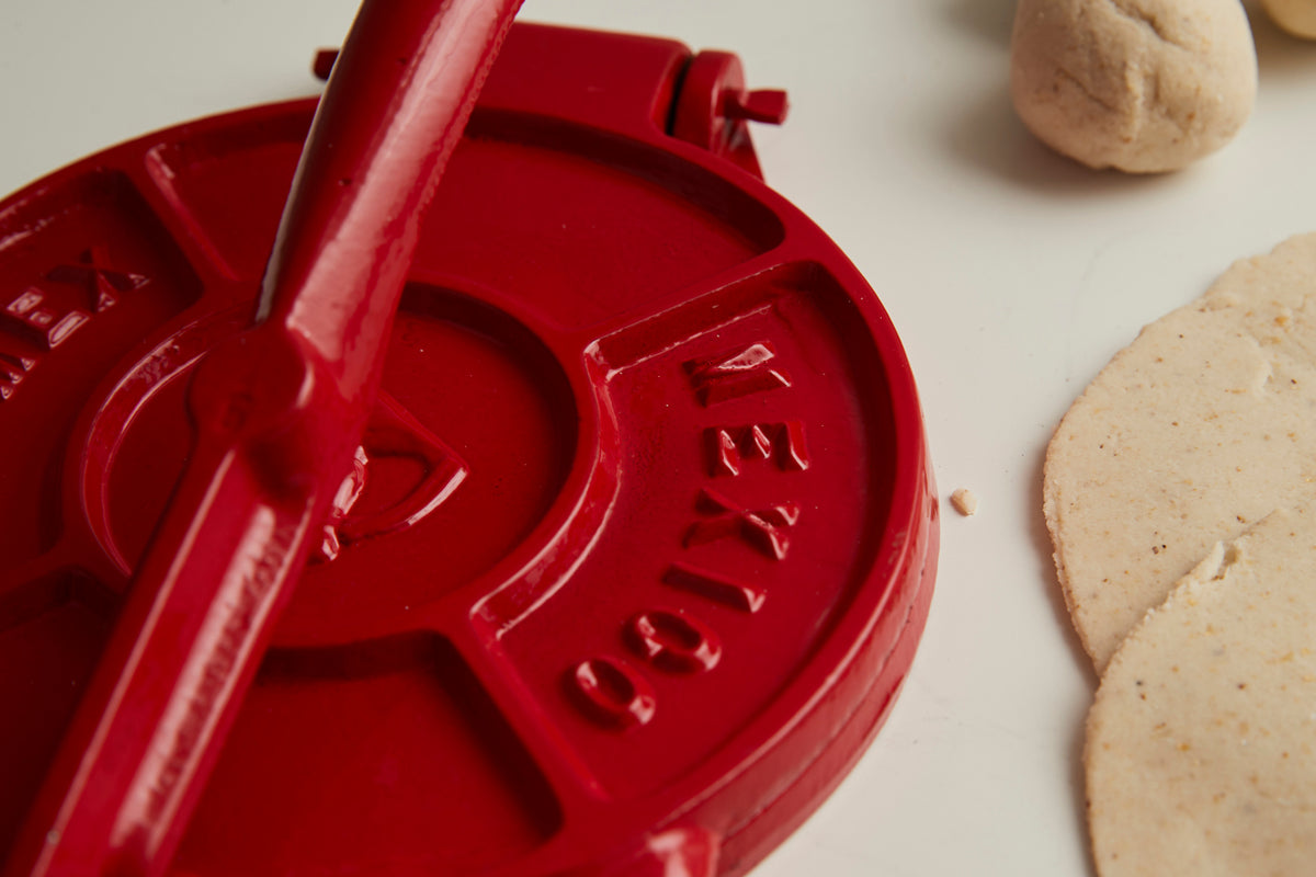 Red cast iron tortilla press with handle shown open, alongside cooked tortillas and dough ball.