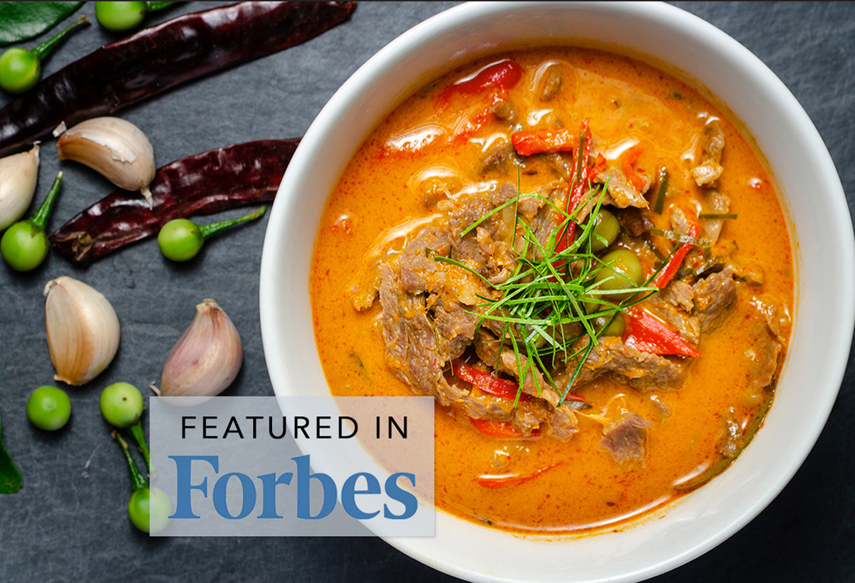 White bowl of orange Panang curry with beef, garnished with green onions and red chilies, surrounded by garlic, dried chilies