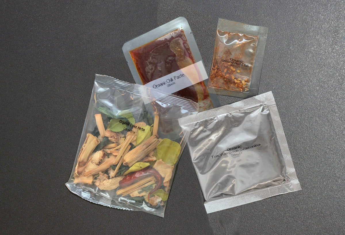 Four sealed packets containing red soup base, dried spices, fresh vegetables, and powder ingredient for tom yum soup.
