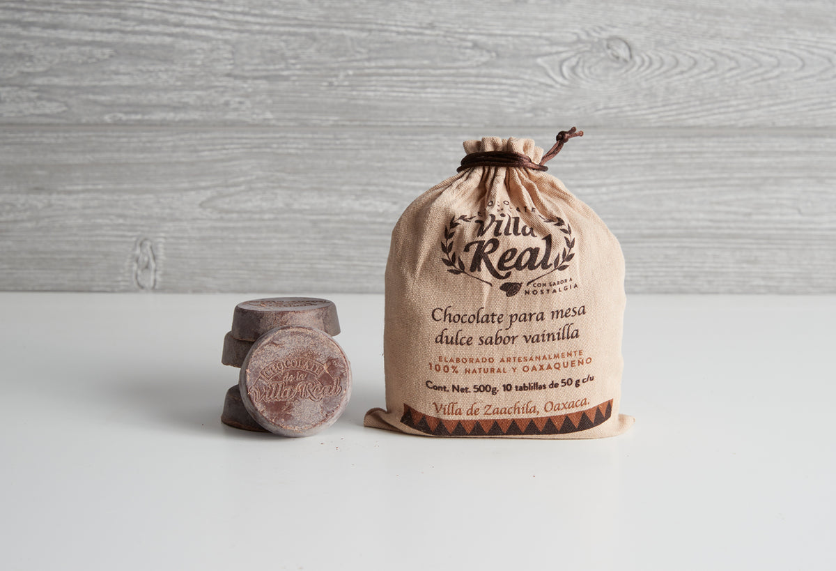 Villa Real Mexican hot chocolate tablets and cloth bag with product information and vanilla flavor text on wooden background.