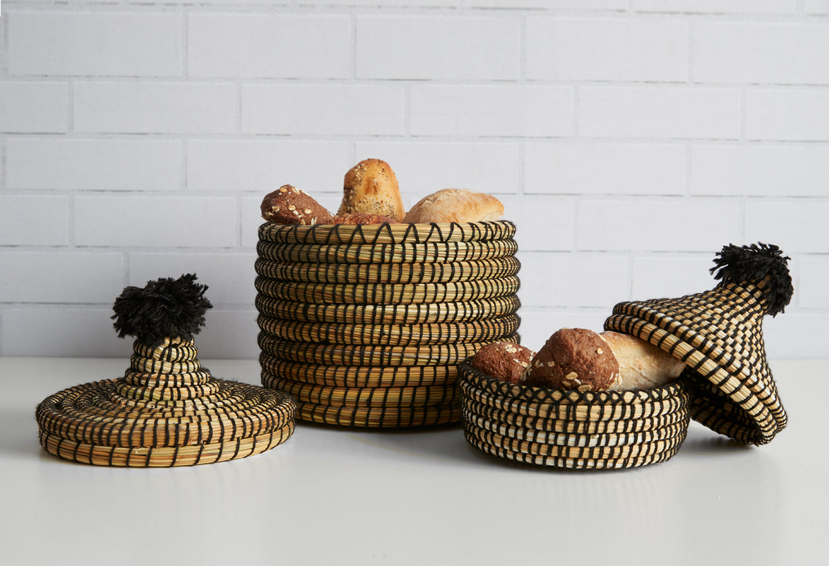 Two woven bread baskets with black tassels, one containing loaves of bread, displayed against a white brick wall.