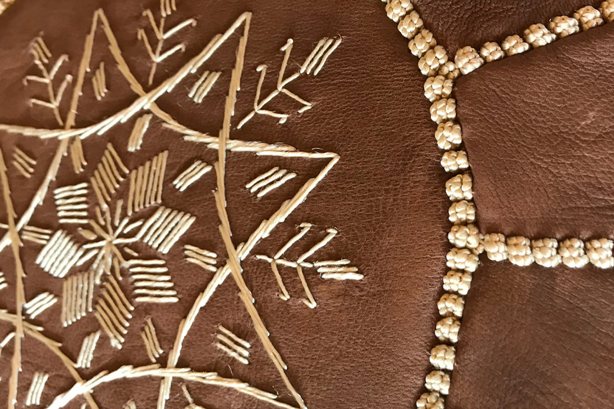 Brown leather pouf with tan embroidered floral branches and beaded trim detail.
