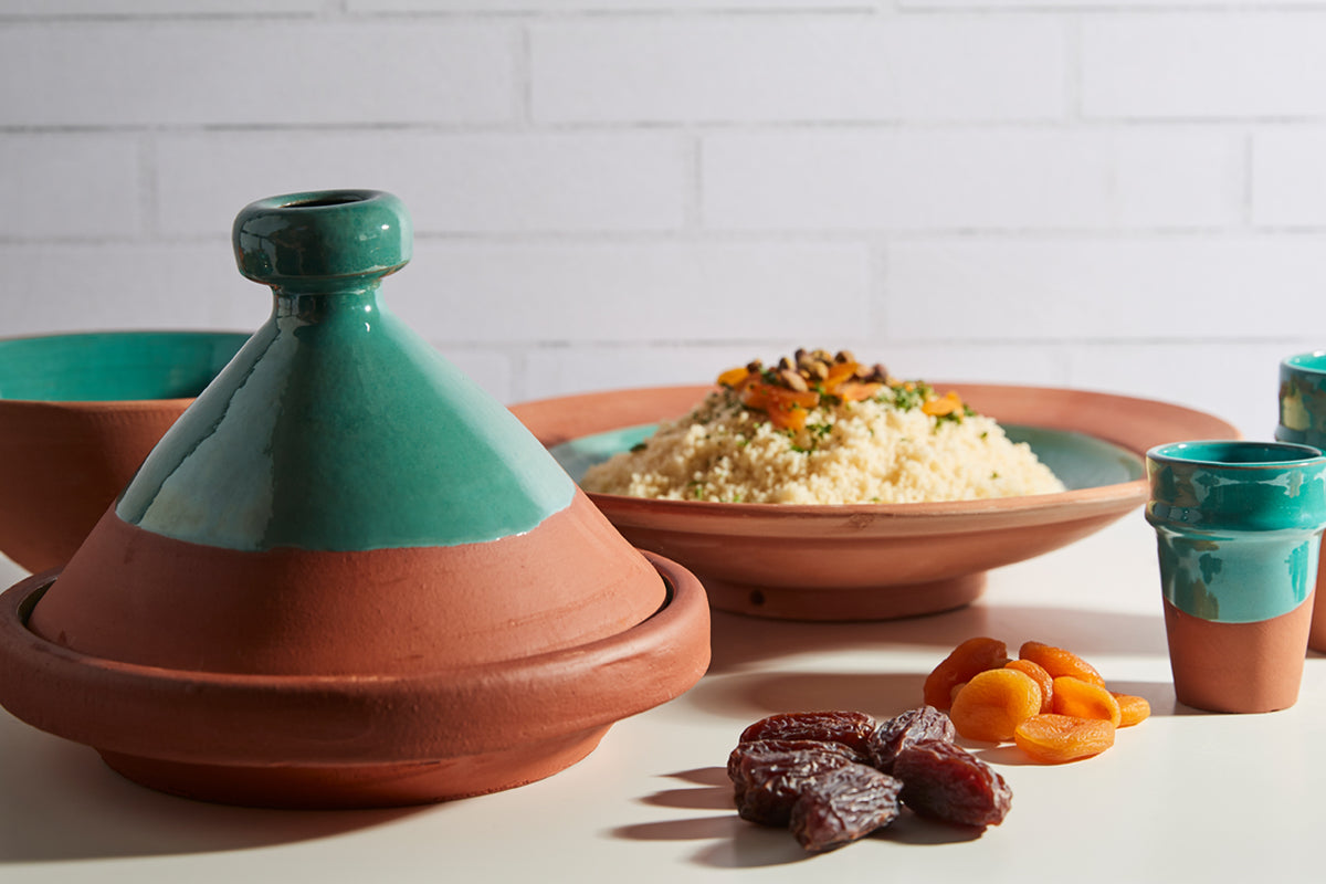 Terracotta tagine with green cone-shaped lid, displayed with couscous, dried apricots, and dates on white surface.