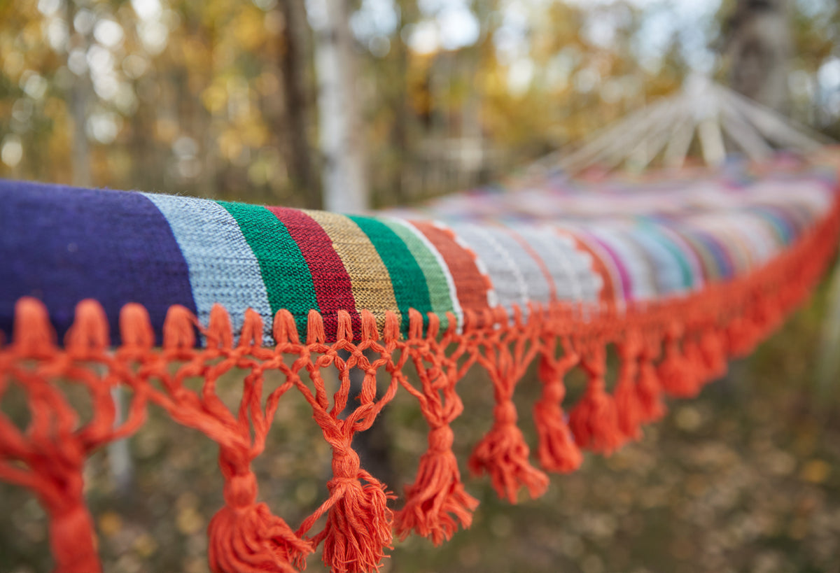 Woven hammock with multicolored horizontal stripes and red knotted fringe trim, displayed outdoors.