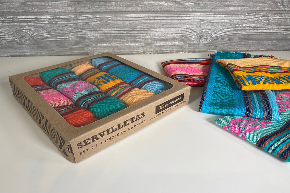 Colorful striped Mexican napkins in cardboard box labeled "Servilletas," displayed on white surface with additional folded na