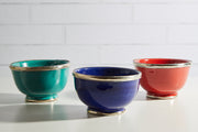 Three glazed bowls in turquoise, navy blue, and coral red with silver-tone metal trim around rims and bases.