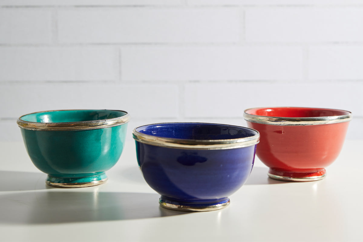 Three glazed bowls in turquoise, navy blue, and coral red with silver-tone metal trim around rims and bases.