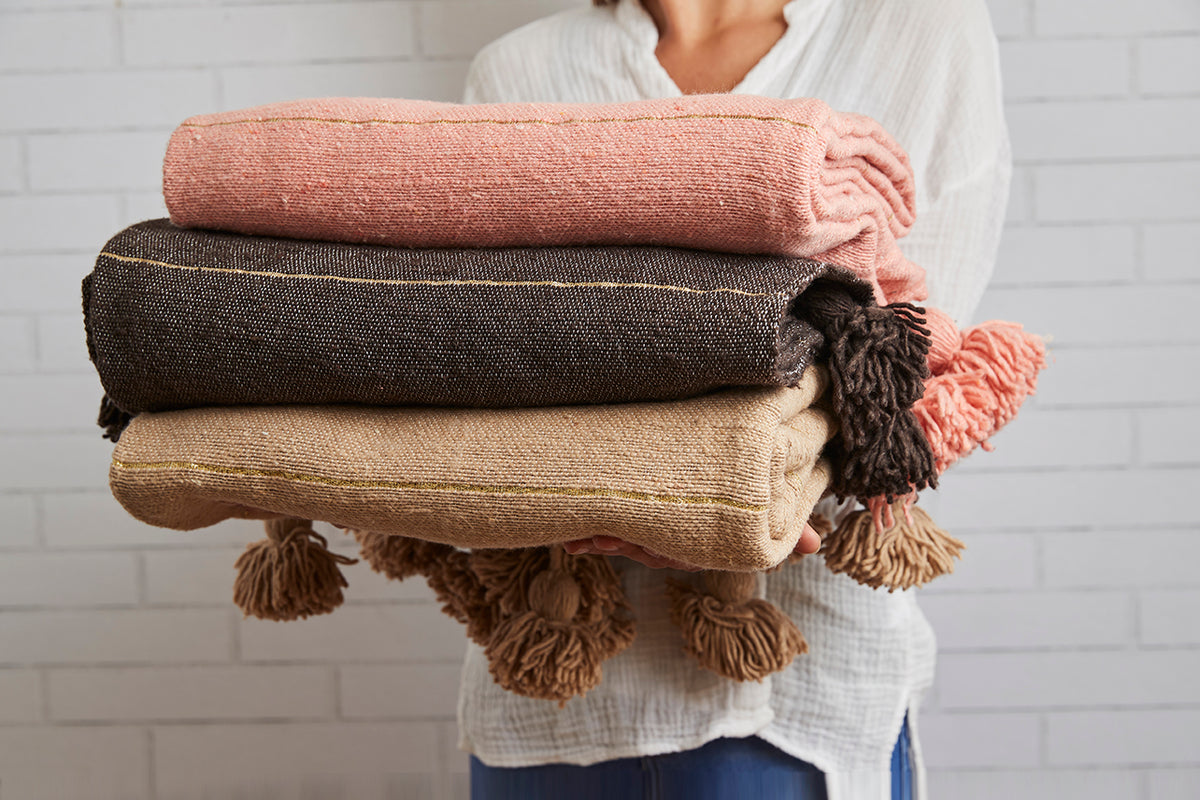 Stack of three folded blankets in pink, charcoal, and beige with tasseled edges held by a person.