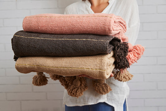 Stack of three folded blankets in pink, charcoal, and beige with tasseled edges held by a person.