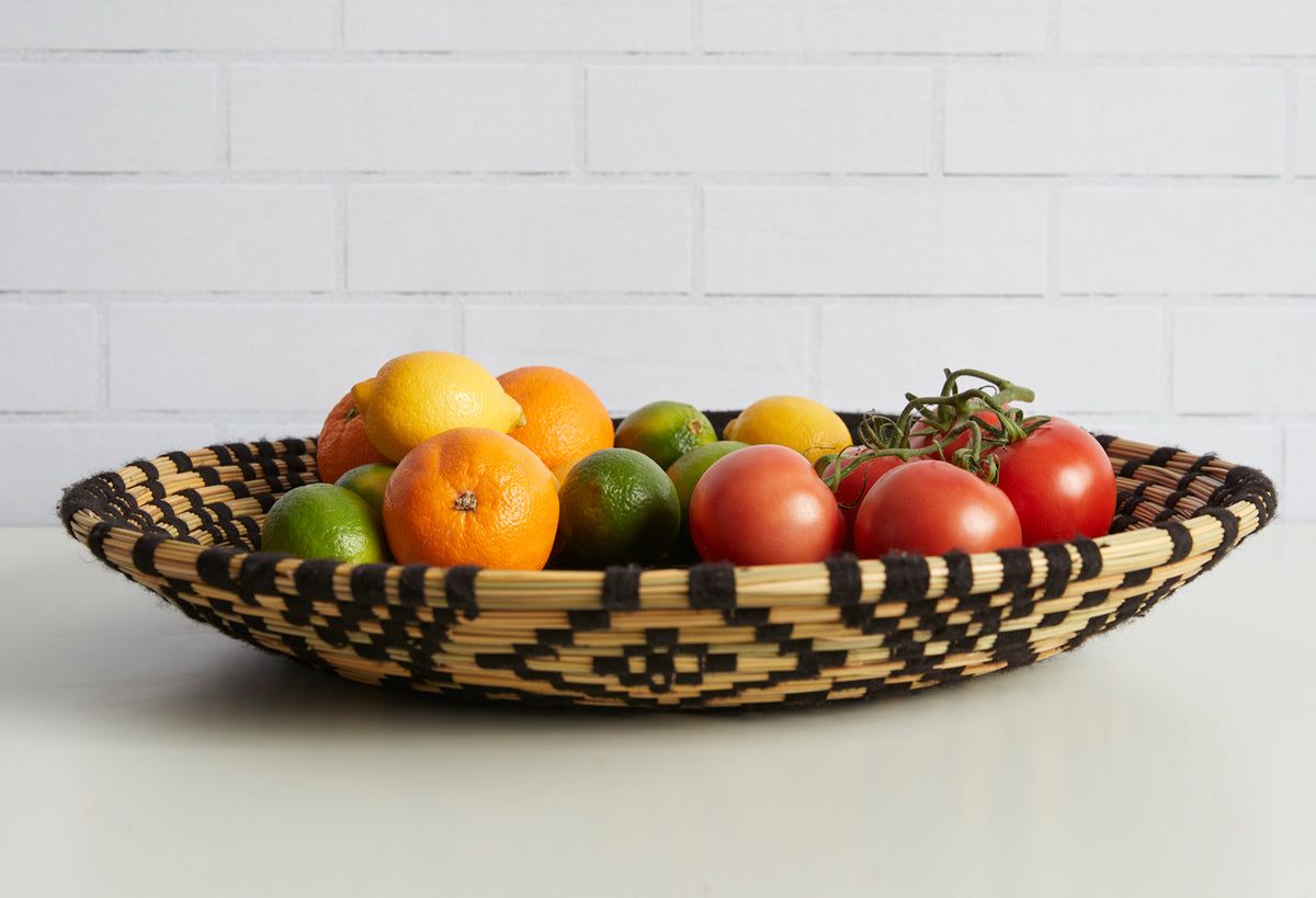 Woven tray with black and tan geometric pattern holding lemons, oranges, limes, tomatoes, and herbs.