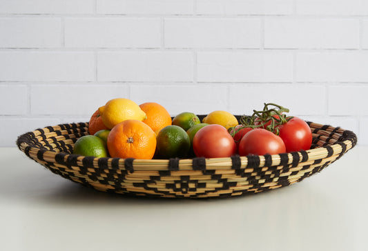Woven tray with black and tan geometric pattern holding lemons, oranges, limes, tomatoes, and herbs.